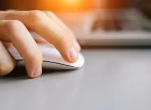 A close-up of a hand using a white computer mouse on a gray desk with a blurred laptop keyboard in the background and warm sunlight shining.