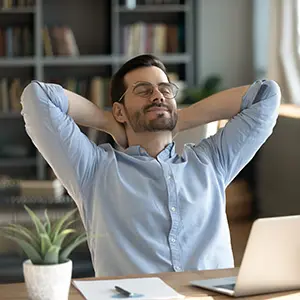A man wearing glasses and a light blue shirt leans back in his chair with his hands behind his head, smiling and looking relaxed at a desk with a laptop, notebook, and potted plant. Bookshelves are in the background.