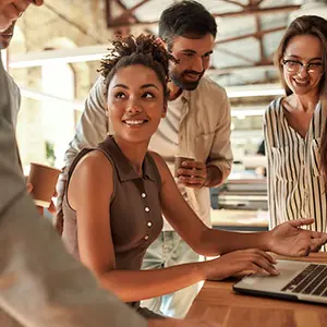 A smiling woman sits at a desk using a laptop while three colleagues stand around her, engaged in conversation in a bright, modern office space.