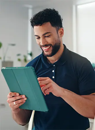 A man with curly hair and a beard, wearing a navy polo shirt, smiles while using a green tablet in a brightly lit indoor space.