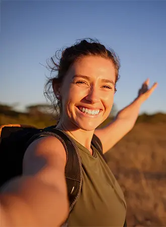 A woman with a backpack smiles brightly and stretches her arm out while taking a selfie outdoors at sunset, with a blurred background of dry grass and trees.