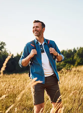 A man wearing a blue shirt, white t-shirt, and brown shorts stands in a grassy field with a backpack, smiling and looking into the distance on a sunny day. Trees are visible in the background.