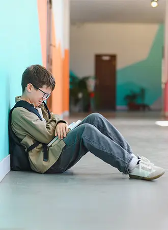 A boy wearing glasses and a backpack sits on the floor against a colorful wall in a hallway, looking down at an open book in his lap.
