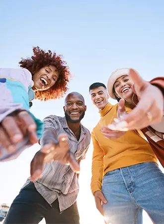 Four young adults stand close together outdoors, smiling and reaching toward the camera with open arms. The sky is clear and blue in the background, creating a vibrant and joyful atmosphere.