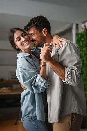 A smiling couple stands close together indoors, embracing and laughing. The woman wears a light denim shirt, while the man wears a light gray button-up shirt. They appear happy and affectionate.