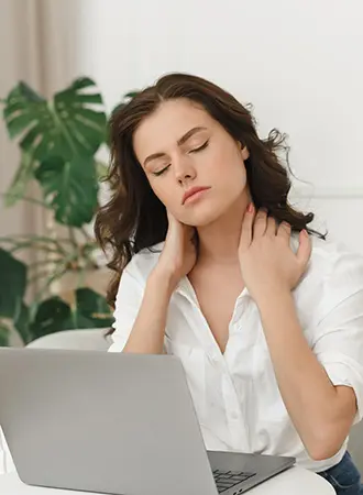 A woman sits at a desk with a laptop, eyes closed, gently massaging her neck with both hands, appearing tired or in discomfort. A large green plant is in the background.