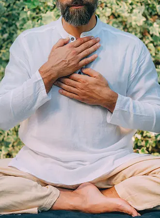 A person with a beard sits cross-legged outdoors, wearing a white shirt and beige pants, with both hands placed on their chest in a peaceful, meditative posture.