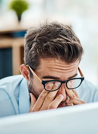 A man wearing glasses rubs his tired eyes while sitting at a desk, suggesting fatigue or eye strain, with a blurred background of an office environment.