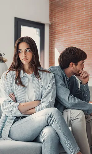 A young woman and man sit back-to-back on a couch, both looking away and appearing upset or in disagreement. The woman has her arms crossed and the man rests his chin on his hand, gazing to the side.