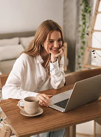 A woman with long hair, wearing a white shirt, sits at a wooden table with a cup of coffee and smiles while looking at a laptop in a cozy, sunlit room.