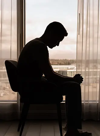 A person sits in a chair indoors, silhouetted against a large window with sheer curtains, head bowed and hands clasped, appearing deep in thought.