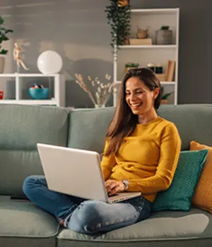 A woman in a yellow sweater sits cross-legged on a green couch, smiling while using a laptop. Behind her are shelves with decorative items and plants in a cozy, well-lit living room.